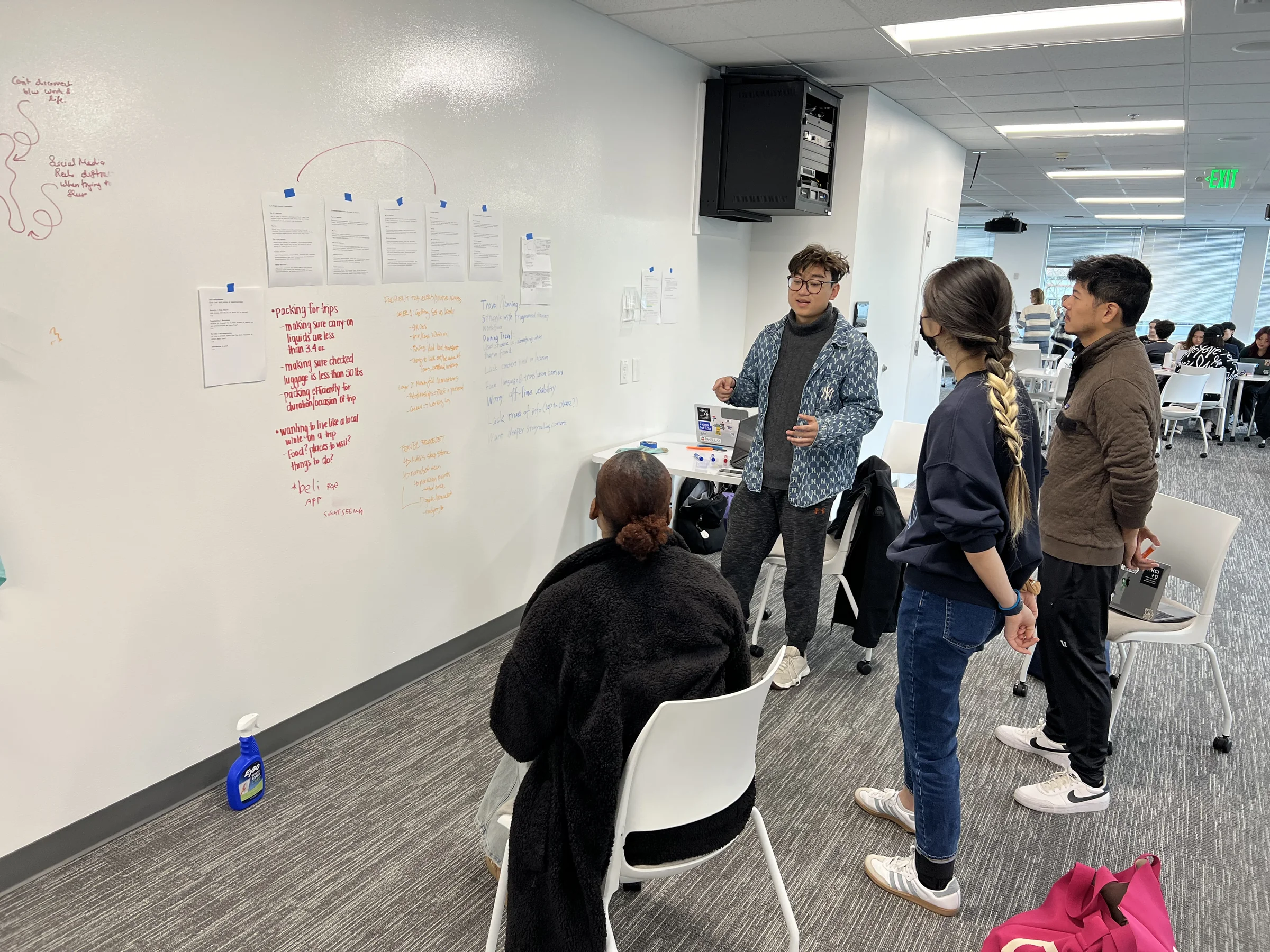 The team at the whiteboard during discovery: interview notes pinned above handwritten clusters on packing, planning, and in-flight discomfort, with Malik talking the group through them
