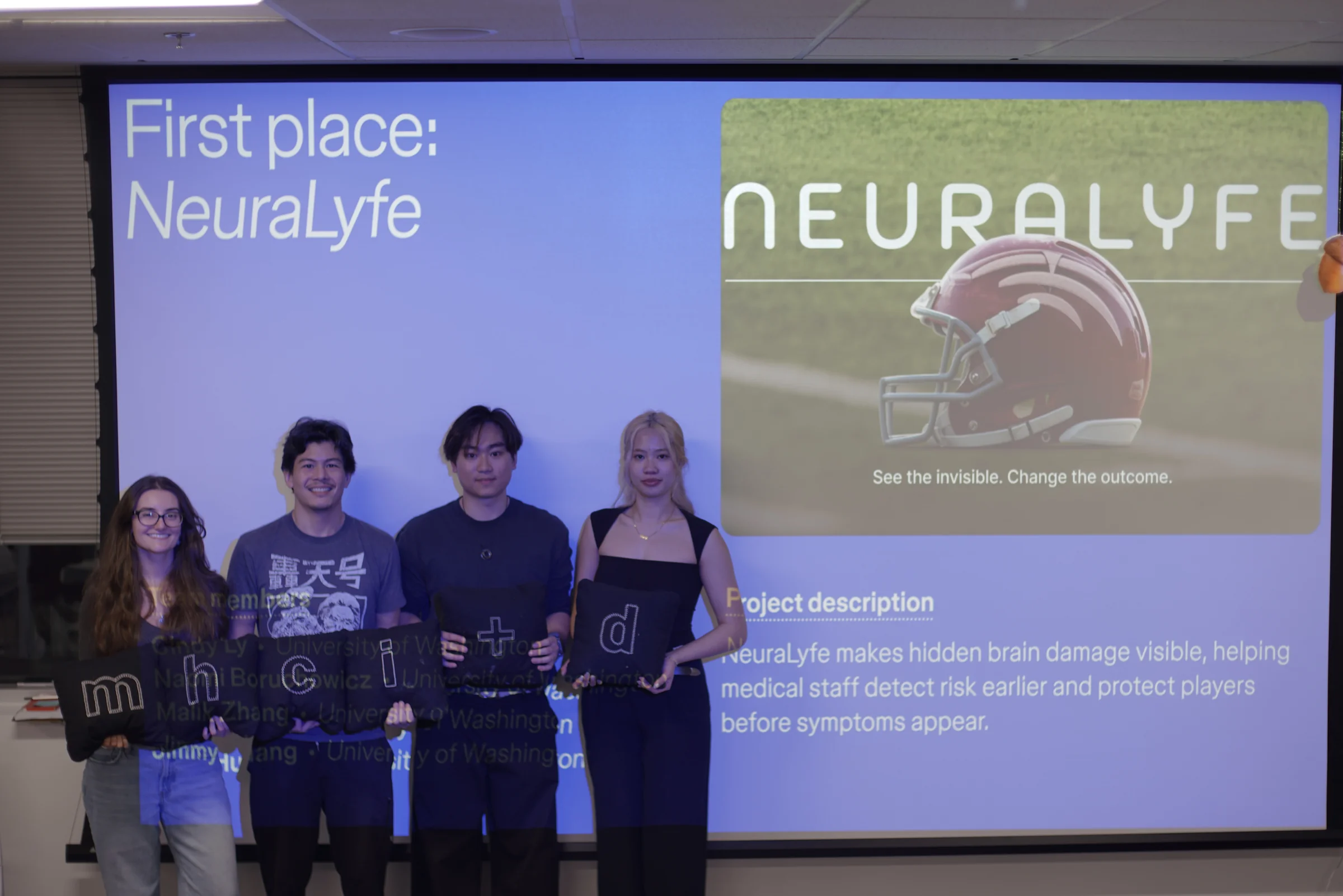 The four NeuraLyfe teammates holding MHCI+D letter cushions in front of the projected First place: NeuraLyfe slide at the FigBuild 2026 awards